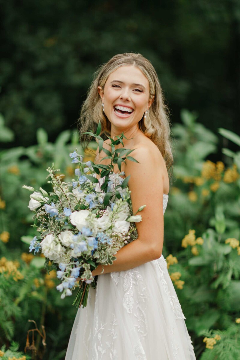 Bride holding a bouquet of white and blue flowers, smiling outdoors.