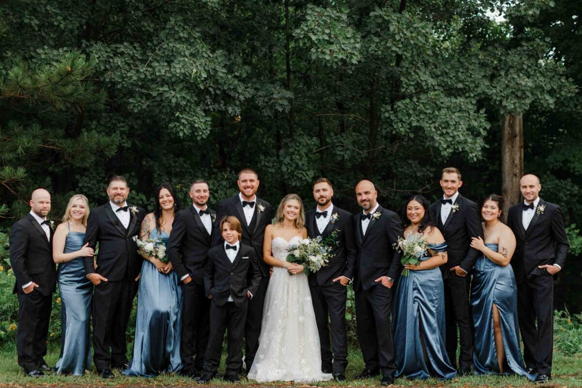 Wedding party standing outdoors near a lake with lush trees in the background.