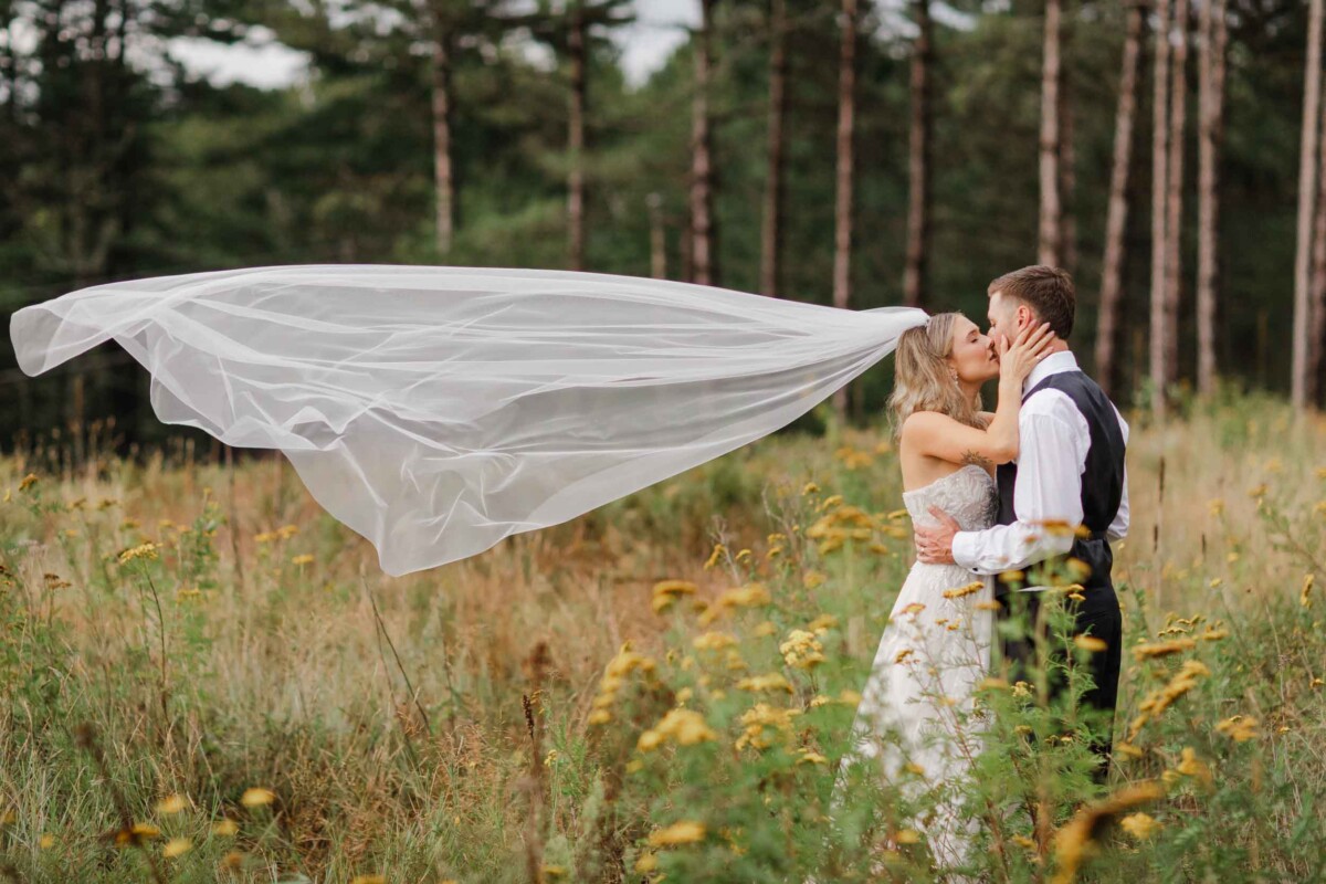 Bride and groom sharing a kiss outdoors with flowing veil in a grassy field.