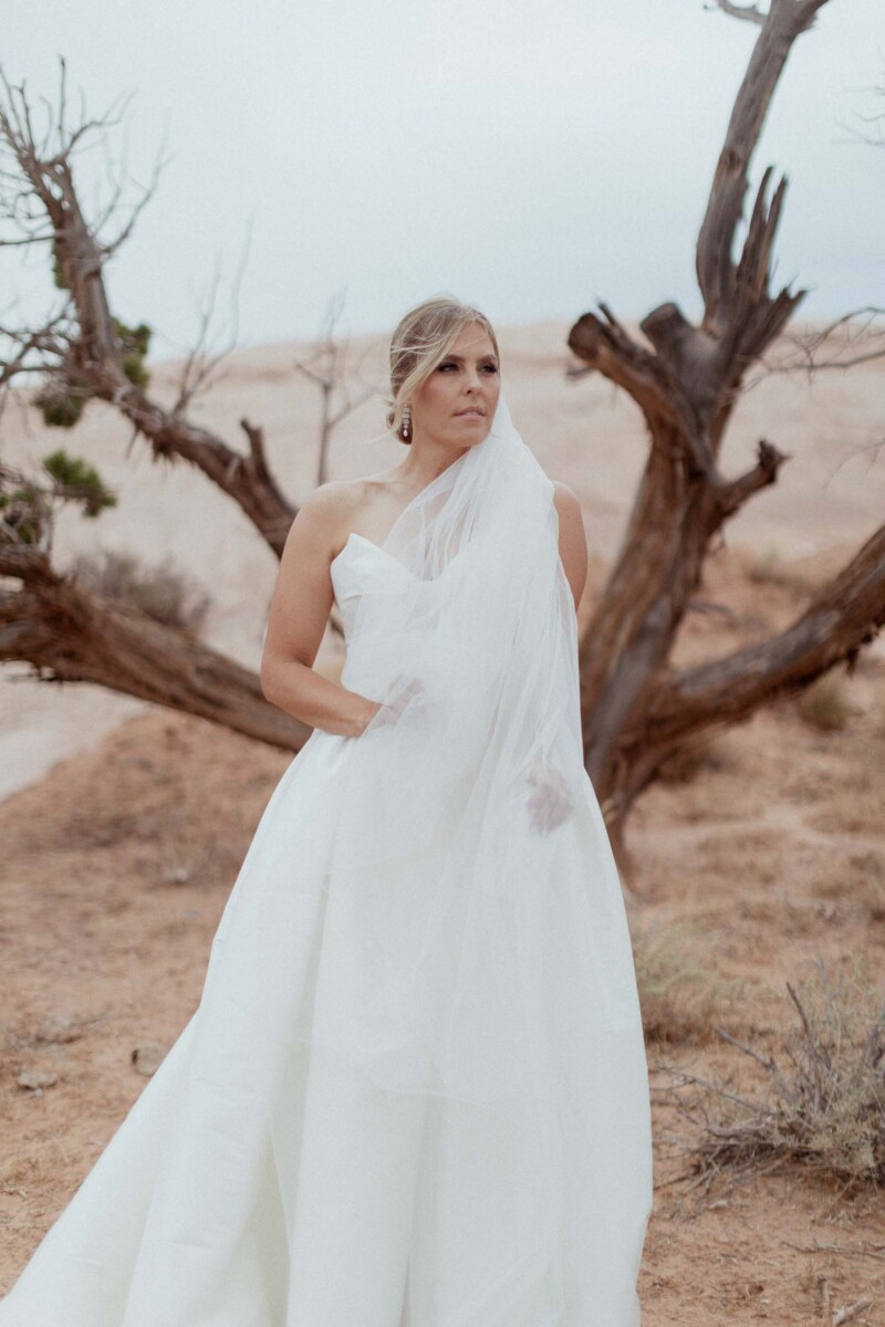 Bride in white dress standing near desert tree in Moab Utah.
