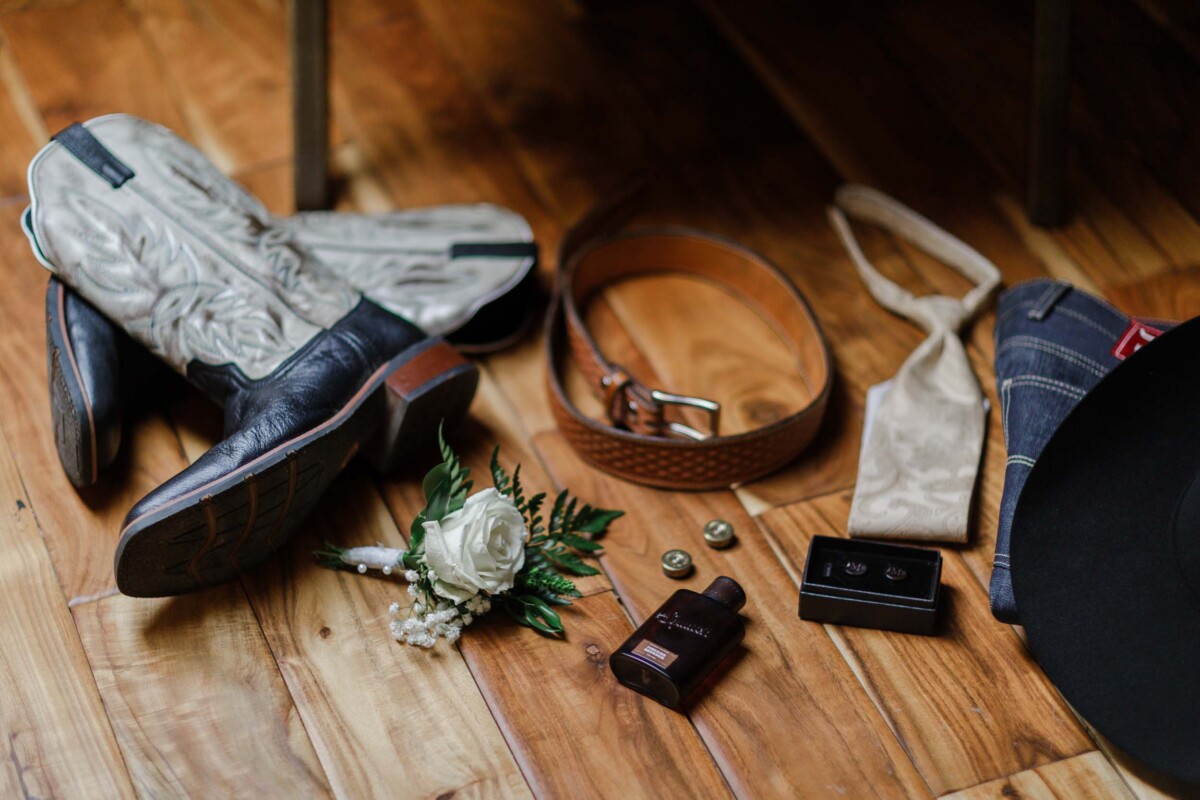 Elegant wedding accessories including shoes, belt, boutonniere, and camera on wooden floor.