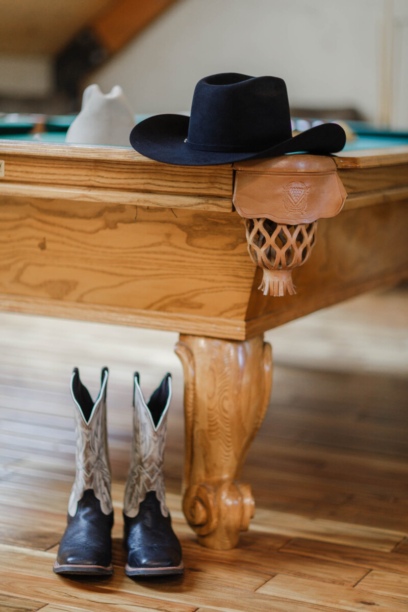 Close-up of cowboy boots and a black hat hanging on a wooden table edge.