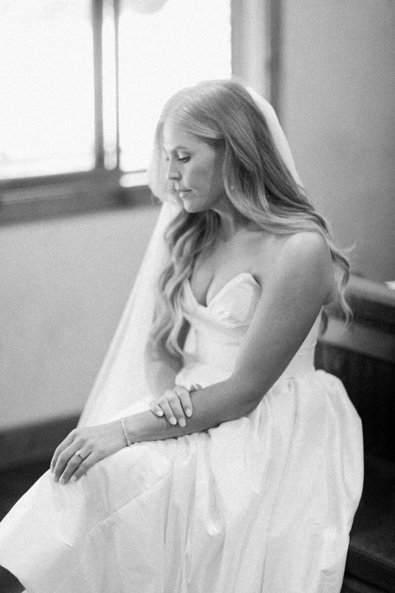 Bride sitting indoors in her wedding dress, looking down thoughtfully.