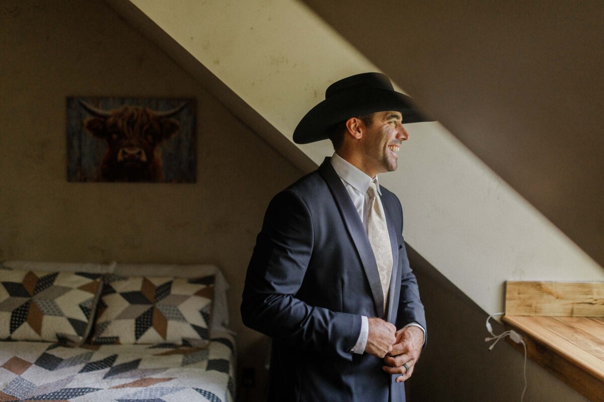 Groom in a suit and cowboy hat smiling indoors during a rustic Utah wedding.