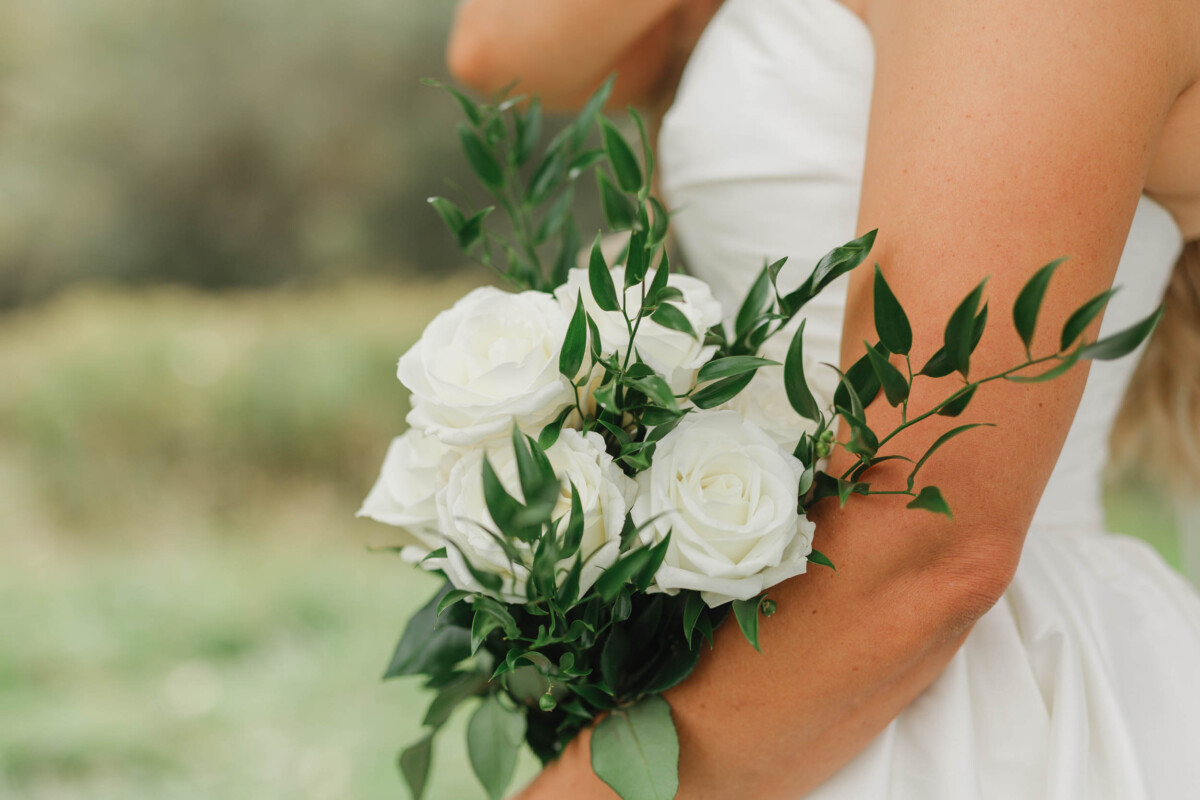 Bride holding a white rose bouquet with greenery at a rural wedding.