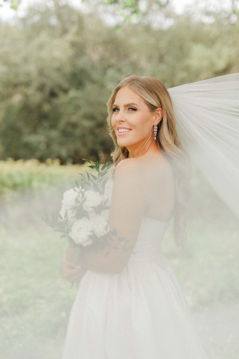 Bride holding bouquet with flowing veil in natural outdoor setting.