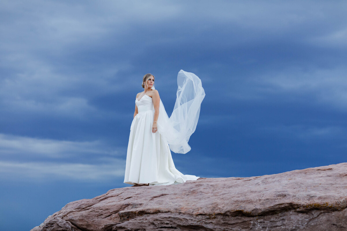 Bride standing on a rocky ledge with a flowing veil against a moody sky.