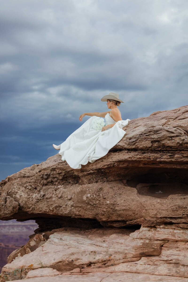 Bride sitting on rock formation in Moab Utah with dramatic sky.