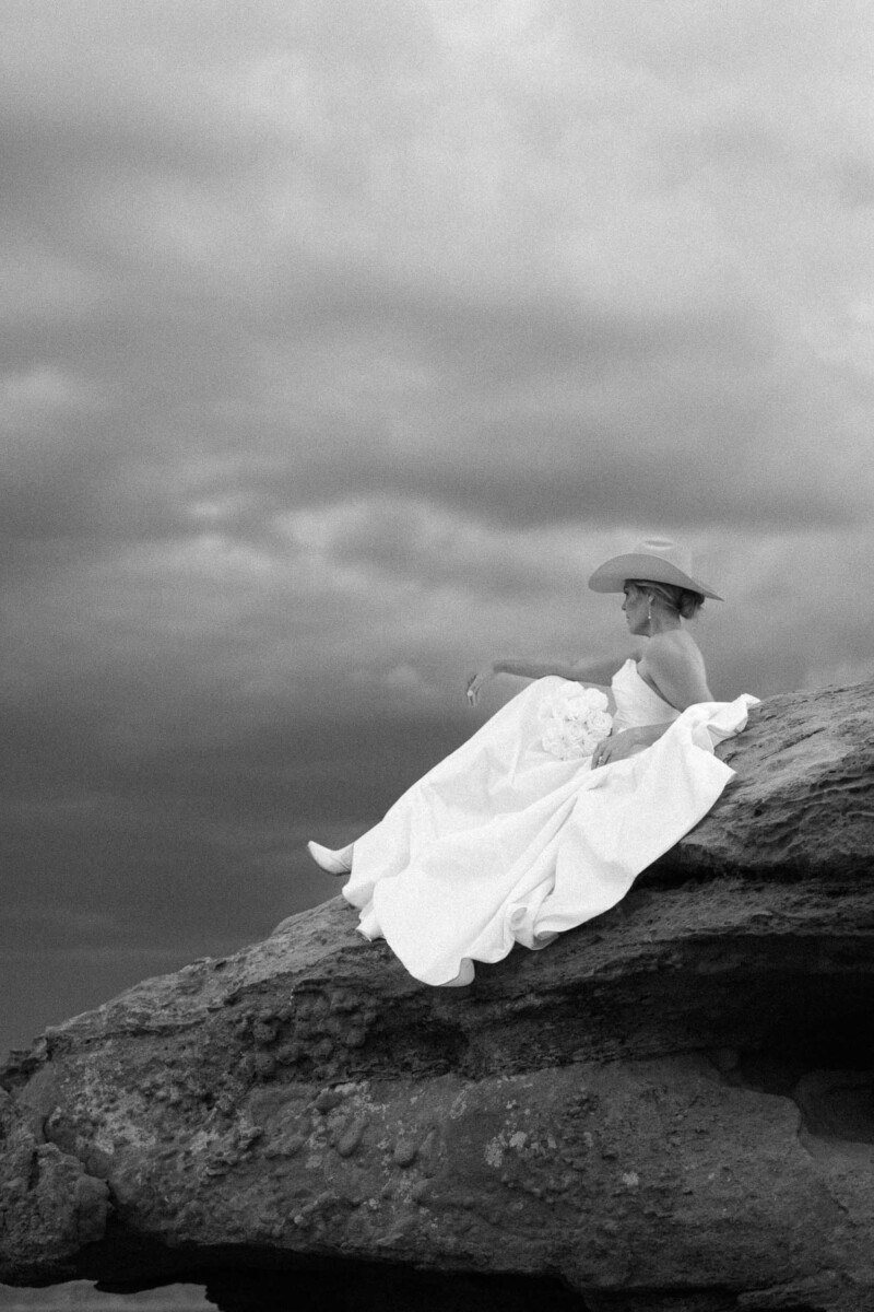 Elegant bride sitting on rock in Moab, Utah, wearing a flowing wedding gown and wide-brim hat.