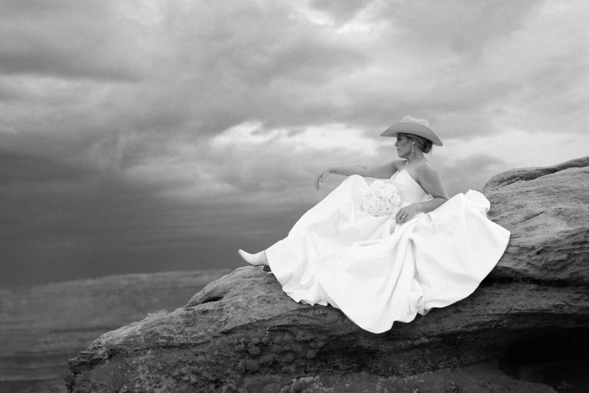 Bridal portrait of a woman in a white dress and hat sitting on a rock in Moab, Utah, with dramatic c.