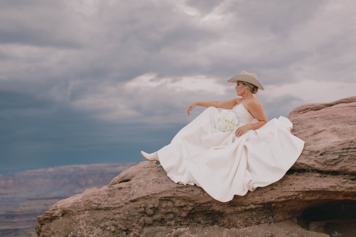 Bride in white wedding dress and cowboy hat relaxing on rock formation in Moab Utah.
