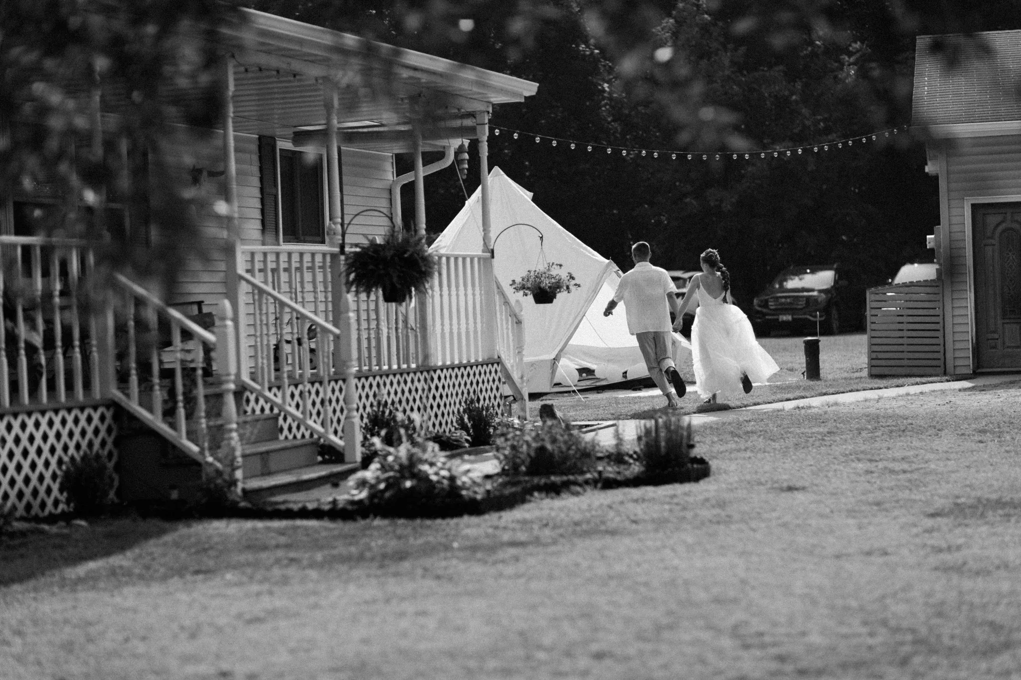 Black and white photo of bride and groom running past white farmhouse cottage at Wisconsin Dells backyard wedding
