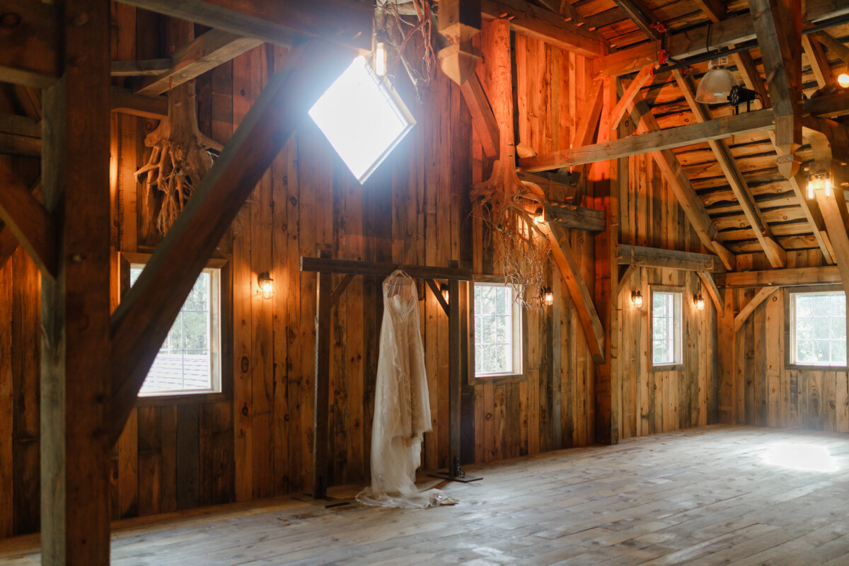 Lace wedding gown hanging in the timber-frame loft of The Swan Barn Door, lit by a diamond window