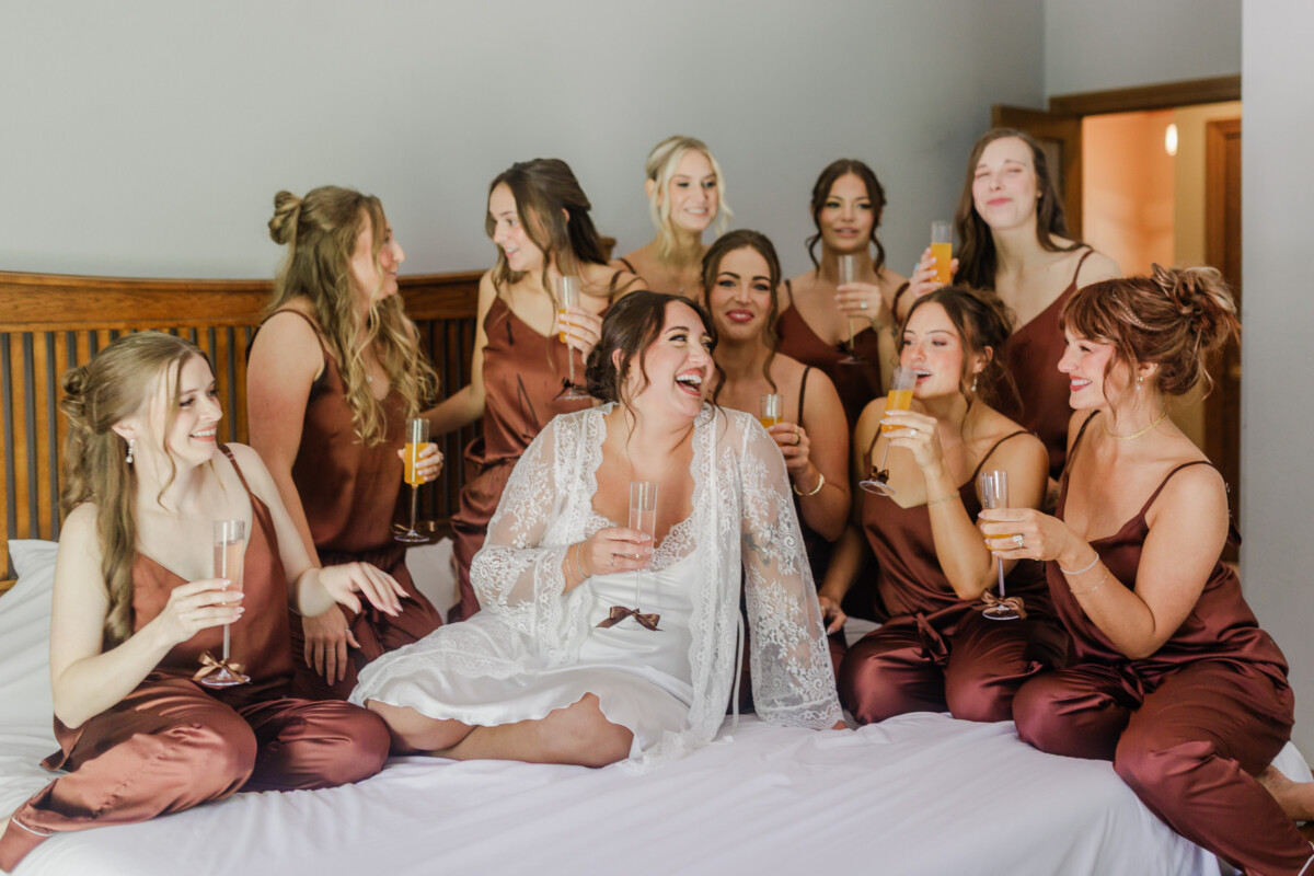 Bride in a lace robe laughing with bridesmaids in rust satin pajamas during a mimosa toast