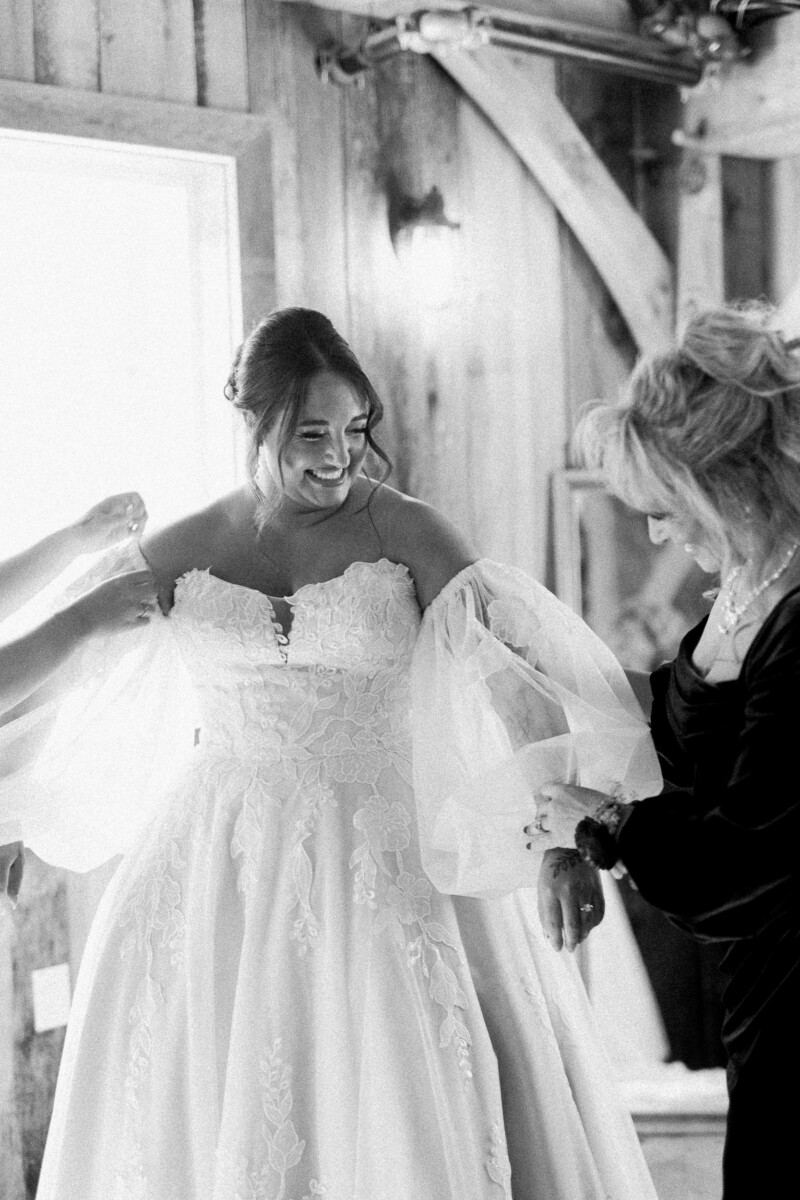 Black and white photo of the bride smiling as she is helped into her off-the-shoulder lace gown