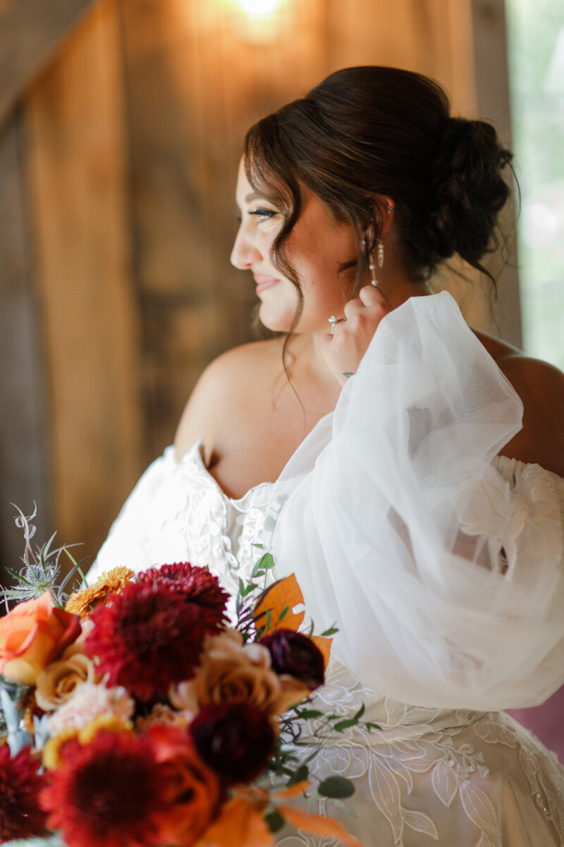 Bride in profile with a puff-sleeve lace gown and fall bouquet beside a barn window
