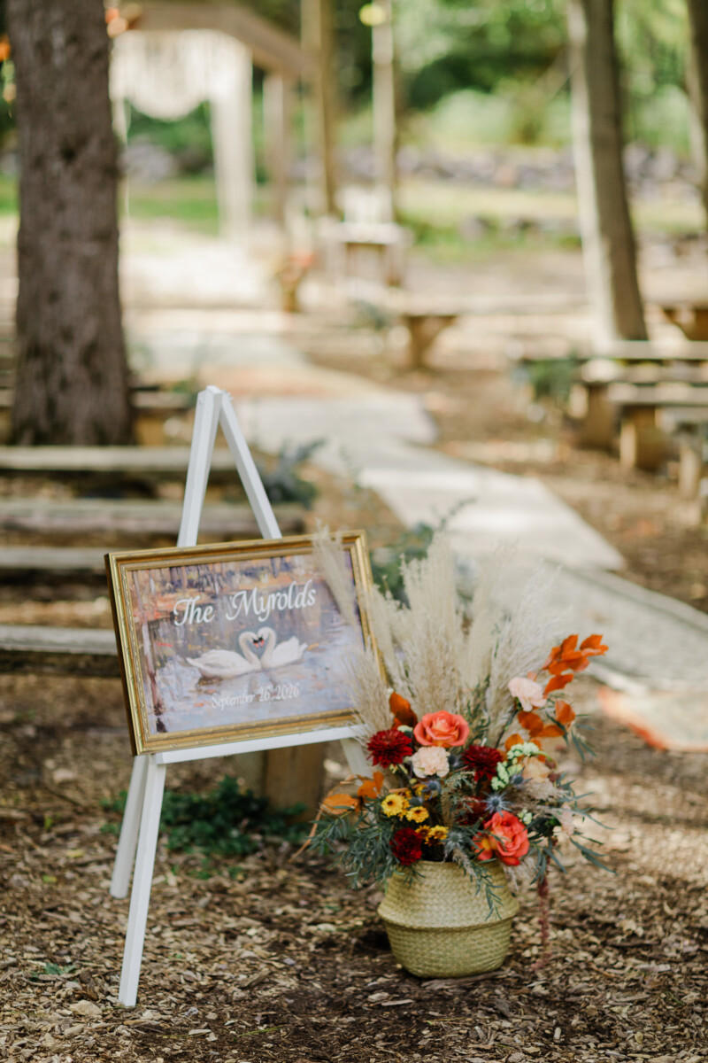 Swan-painting welcome sign with pampas and dahlias at the wooded ceremony site at The Swan Barn Door