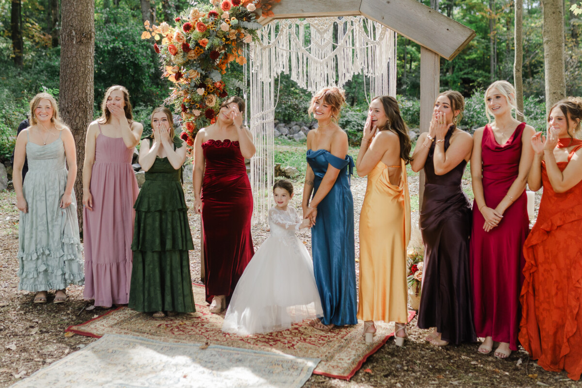 Bridesmaids in jewel-tone gowns react with tears and smiles beneath a macramé ceremony arch