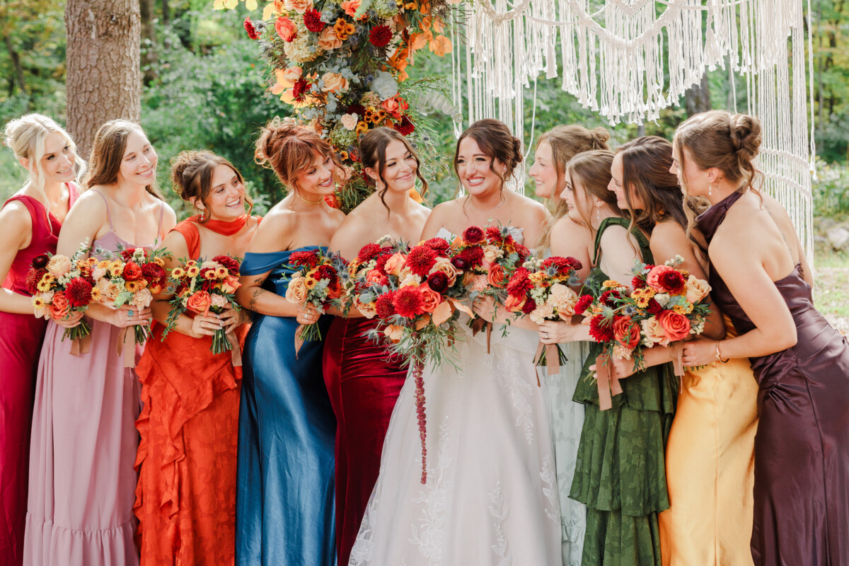 Bride laughing with bridesmaids in mismatched jewel-tone gowns under the macramé arch