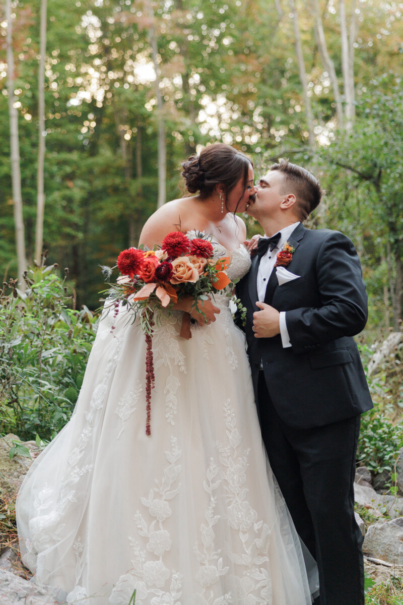 Bride and groom kissing in the forest at sunset, her bouquet at her side